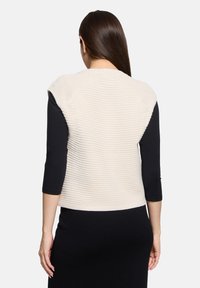 Veste beige texturée avec un design côtelé, manches courtes et encolure ronde, portée sur un haut noir à manches longues.