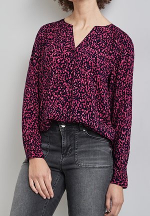 Femme portant un chemisier à manches longues rose et noir à motifs, rentré dans un jean noir délavé taille haute, debout devant un fond uni.