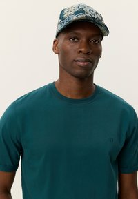 Τυρκουάζ βαμβακερό μπλουζάκι με κοντά μανίκια και στρογγυλή λαιμόκοψη, με ένα μικρό κεντημένο λογότυπο. Φορεμένο με καπέλο με σχέδιο σε μπλε αποχρώσεις.