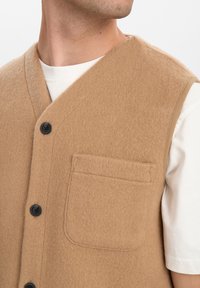 Gilet di lana beige con scollo a V, bottoni frontali e una singola tasca sul petto. Tessuto liscio e leggermente texturizzato, con bottoni neri e leggere spalle accentuate.