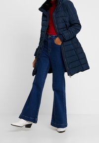 Abrigo puffer azul marino con cierre de cremallera, combinado con unos vaqueros de tiro alto y acampanados en azul oscuro y botas de tobillo blancas con tacones cuadrados.