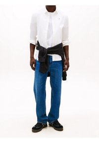 Bílá košile s knoflíky přetažená přes bílý tričko, v kombinaci s modrými džínami a černými teniskami, s tmavým svetrem uvázaným kolem pasu.