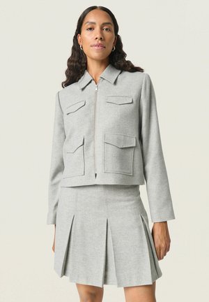 Grijze cropped jack met twee opgestikte zakken, zilveren rits en lange mouwen, gecombineerd met een bijpassende geplooide minirok.