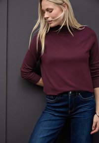 Bordeaux-farvet, tætsiddende sweater med høj krave. Parret med mørkeblå jeans med klassisk fem-lommer design og subtil kontrastsyning.