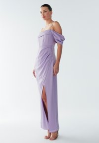 Femme debout dans une robe lavande épaules dénudées avec une fente haute jusqu'à la cuisse et des talons à brides transparentes, sur un fond blanc uni.