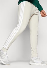 Šviesiai rudos spalvos sportiniai kelnių su baltais trimis dryžiais šonuose. Turi siaurėjantį siluetą ir pūkuotas rankoves. Pagaminta iš minkštos, tekstūruotos medžiagos.