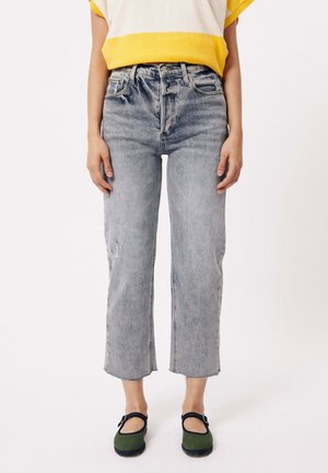 Verblasste blaue hochtaillierte Jeans mit geradem Bein, mit abgetragenen Details, fransigem Saum, kombiniert mit grünen Schuhen und einem top mit gelben Akzenten.