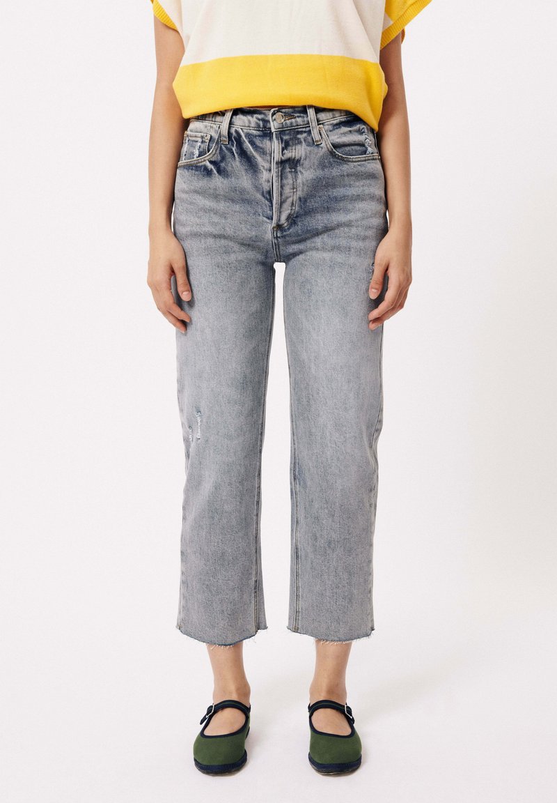 Vervaagde blauwe high-waisted jeans met een rechte pijp, verweringdetails, rafelige zoom, gecombineerd met groene schoenen en een top met gele accenten.