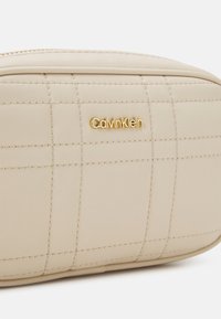 Béžová prošívaná kosmetická taška z umělé kůže se zlatým logem "CalvinKlein", s hladkou texturou a jemnými detaily prošití.