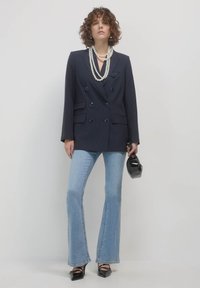 Donna con capelli ricci che indossa un blazer a righe blu navy, collane di perle sovrapposte, jeans a campana chiari, tacchi neri e tiene una piccola borsa nera.
