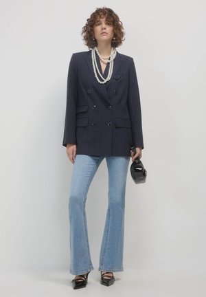 Donna con capelli ricci che indossa un blazer a righe blu navy, collane di perle sovrapposte, jeans a campana chiari, tacchi neri e tiene una piccola borsa nera.