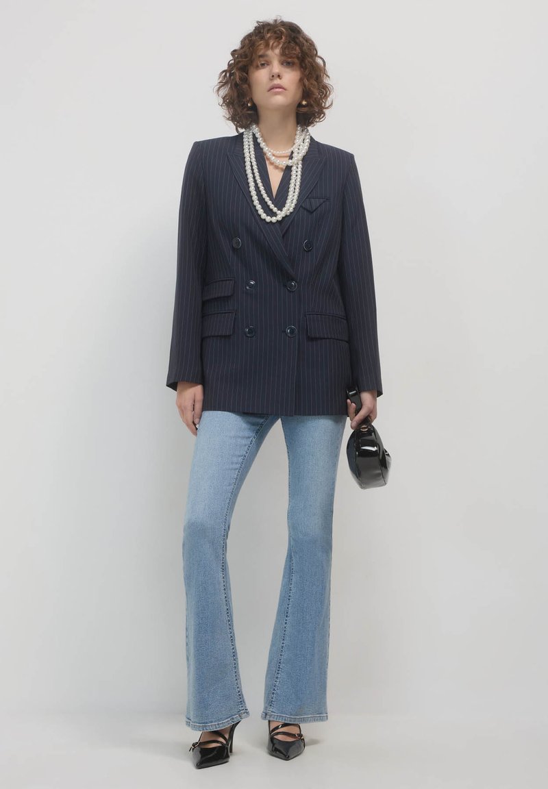 Donna con capelli ricci che indossa un blazer a righe blu navy, collane di perle sovrapposte, jeans a campana chiari, tacchi neri e tiene una piccola borsa nera.