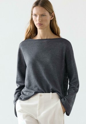 Kvinde med langt hår iført en mørkegrå langærmet sweater og lysebeige højttaljede bukser, hænderne i lommerne, neutral baggrund.