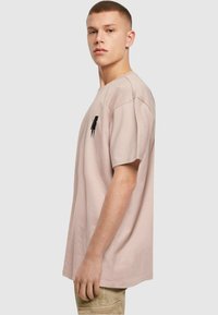 Lys pink bomulds T-shirt med korte ærmer, afslappet pasform og et sort abstrakt design på brystet. Glat tekstur, simpelt design.