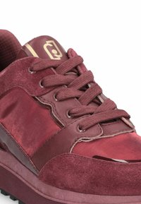 Gros plan sur une sneaker bordeaux avec des panneaux en daim et en cuir, des lacets épais et un logo doré sur la languette, le tout sur fond blanc.