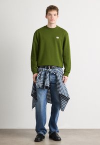 Sweatshirt verde com um pequeno logótipo em patch, camisa xadrez azul atada à cintura, jeans azuis e sapatos pretos num fundo de superfície lisa.