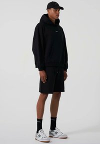 Svarte hettegenser med løs passform og en frontlomme, kombinert med sorte shorts. Tilbehøret inkluderer en svart caps og grå-og-hvite joggesko.