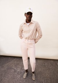 Chemise à rayures beige avec boutons, T-shirt blanc, jean rose pâle et chaussures gris slip-on. Le mannequin porte une casquette blanche avec un design bleu.