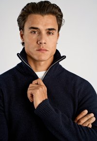 Marineblauer Pullover mit Reißverschluss am Hals, geripptem Kragen und Bündchen, aus weichem, strukturiertem Stoff. Modell hält den Reißverschluss mit einer Hand, die Arme sind verschränkt.