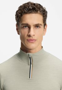 Jasnobeżowa bluzka z kołnierzykiem na zamek, wykonana z teksturowanego materiału. Zamek błyskawiczny zdobiony czarnymi i brązowymi akcentami. Model ma wysoki kołnierz i dopasowany krój.