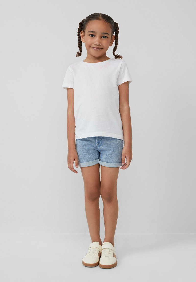 Witte t-shirt met korte mouwen en kleine roze hartdetails, gecombineerd met opgerolde denimshorts en crèmekleurige sneakers met bruine accenten.