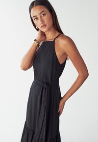 Čierne šaty s výstrihom za krk, priliehavým telom a volánovým lemom. Obsahujú viazaný detail na páse, sú vyrobené z hladkej látky s jemnou textúrou.