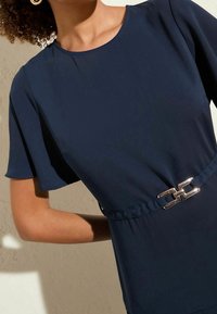 Námořnické modré šaty s krátkými, rozšířenými rukávy a stříbrným řetízkovým páskem kolem pasu, vyrobené z hladkého materiálu s mírným leskem.