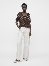 Femme portant un chemisier marron transparent à pois avec des volants, un pantalon blanc à jambes larges et des chaussures blanches à talons bas, debout avec une main dans la poche.