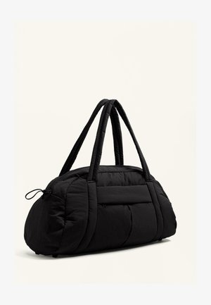 Črn potovalni nahrbtnik z quiltanim dizajnom, opremljen z dvema oblazinjenima ročajema in zadrgo. Material se zdi mehak in teksturiran.