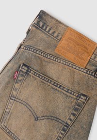 Τζιν σε ανοιχτό μπλε χρώμα με ένα ορθογώνιο δερμάτινο ταμπελάκι στη πίσω μέση, με εμφανείς ραφές και μια κόκκινη ετικέτα στην πλαϊνή τσέπη.