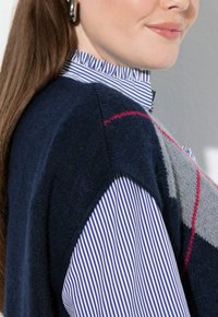 Žena nosící stříbrné kruhové náušnice, modro-bílé pruhované košili s nabíraným límcem a námořnickou modrou a šedou svetrovou mikinu s červenými pruhy.