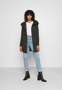 Manteau capuchon vert foncé avec des boutons, porté sur un haut blanc et un jean bleu avec ourlets, associé à des bottines à talons noires. Texture lisse.