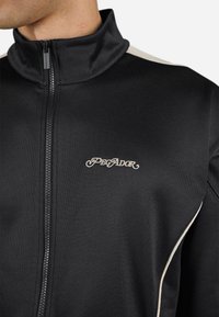 Chaqueta negra con cremallera y cuello alto, con ribetes en color crema a lo largo de los lados y un pequeño logotipo bordado "PEɢADOR" en el pecho.