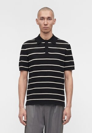 Mężczyzna w czarnej koszulce polo z krótkim rękawem z poziomymi białymi paskami i szarymi spodniami, stojący na jednolitym białym tle.