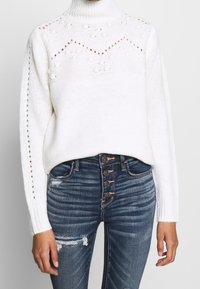 Vit stickad tröja med hög krage, har strukturerade mönster och små bollar; kombinerat med mörkblå slitna högmidjade jeans.