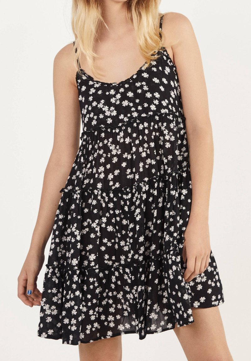 Schwarzes Blumen-Kleid mit weißen Blumen, Spaghettiträgern, Stufendesign und einem gerüschten Saum. Hergestellt aus leichtem Stoff, geeignet für warmes Wetter.