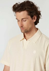 Beige polo tričko s krátkým rukávem, vyrobené z měkké tkaniny, s jemnou nášivkou loga na levé části hrudníku.