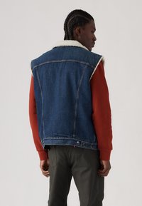 Denimvest med pelskrave, blått stoff, uten ermer, og knapper på baksiden, brukt over en rød hettegenser og olivenfargede bukser.