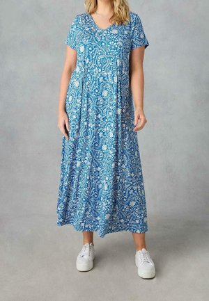 Kvinde iført en blå blomster maxi kjole med korte ærmer og hvide sneakers, stående foran en ensfarvet grå baggrund.