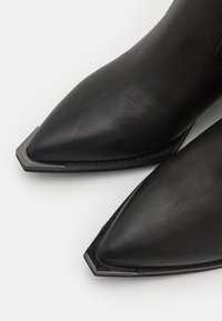 Schwarze Leder-Stiefeletten mit spitzer Zehe und sechseckigem metallischem Akzent an der Zehe. Glatte Textur, elegantes Design und minimale Nähte.