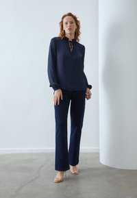 Blusa azul marino de manga larga con cuello de lazo, combinada con vaqueros oscuros de pierna acampanada y tacones abiertos beige. Tela suave con un ligero brillo.