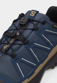 Κοντινή λήψη μπλε και μαύρου παπουτσιού πεζοπορίας με κορδόνια γρήγορης απελευθέρωσης, ανώτερο πλέγμα και λεπτομέρειες μπεζ στη σόλα.