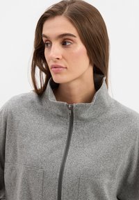 Graue Zip-Jacke mit hohem Kragen, ausgestattet mit einem strukturierten Stoff und zwei aufgesetzten Fronttaschen. Sichtbare, glatte Reißverschlussdetails.