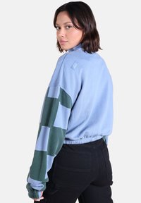 Sweatshirt en polaire bleu clair avec des motifs géométriques verts sur les manches. Caractérisé par un ourlet élastique et une étiquette de marque sur l'épaule. Pantalons noirs visibles.