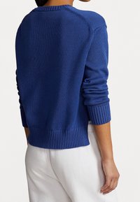 Personne portant un pull bleu en tricot à manches longues avec des poignets et un ourlet côtelés, associé à un pantalon blanc, vue de dos.