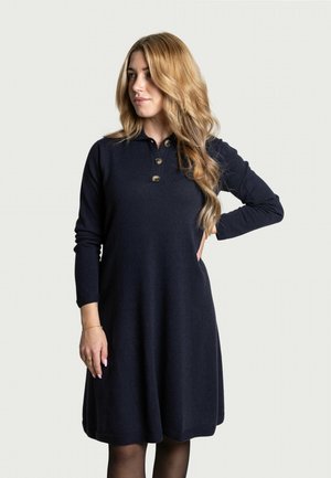 Frau mit langen blonden Haaren, die ein schwarzes Langarmkleid mit drei Knöpfen trägt, steht mit einer Hand auf der Hüfte vor einem schlichten Hintergrund.