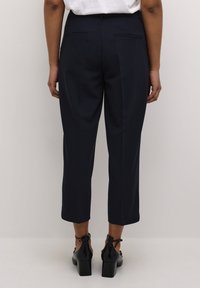 Pantalon bleu marine à coupe ajustée, longueur raccourcie et texture lisse. Comprend deux poches arrière et des coutures nettes. Porté avec des chaussures à talons noires.