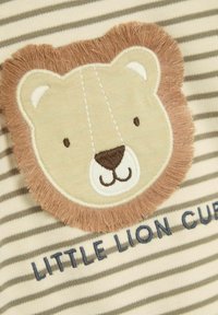 Tissu en coton avec des rayures beiges, comportant un patch brodé de lionceau en marron clair, avec le texte "LITTLE LION CUB" en dessous, en bleu marine.