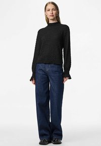 Crni dugih rukava top s teksturom i naboranim izrezom, uparen s širokim tamnoplavim trapericama, nošen s crnim cipelama.