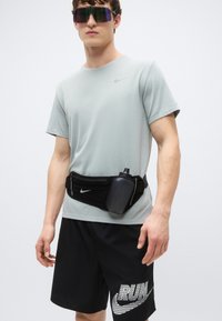 Ljusgrå kortärmad Nike-tröja, svarta shorts med "RUN"-grafik, svart midjeväska med vattenflaska och färgglada solglasögon.